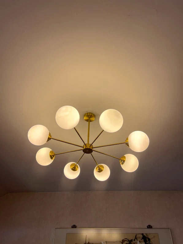 A modern multi-light ceiling fixture with a gold-toned metal frame and eight frosted white glass globes, shown illuminated against a beige wall and ceiling.